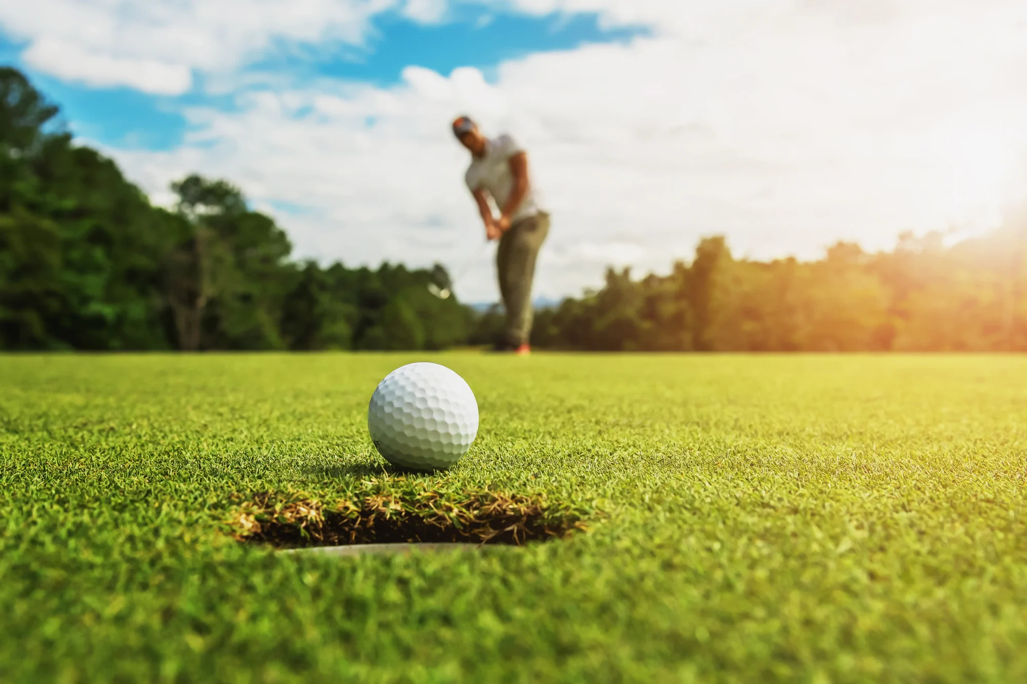 Golf ball rolling toward the cup on a sunlit green as a golfer follows through on a putt