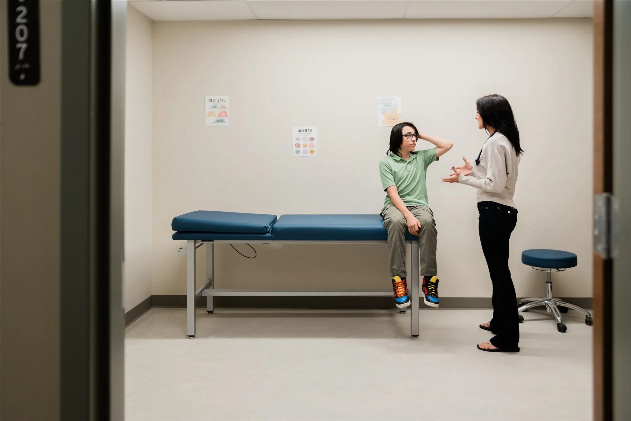 A boy sitting on a medical examination table talks with a standing female doctor in a clinical room.