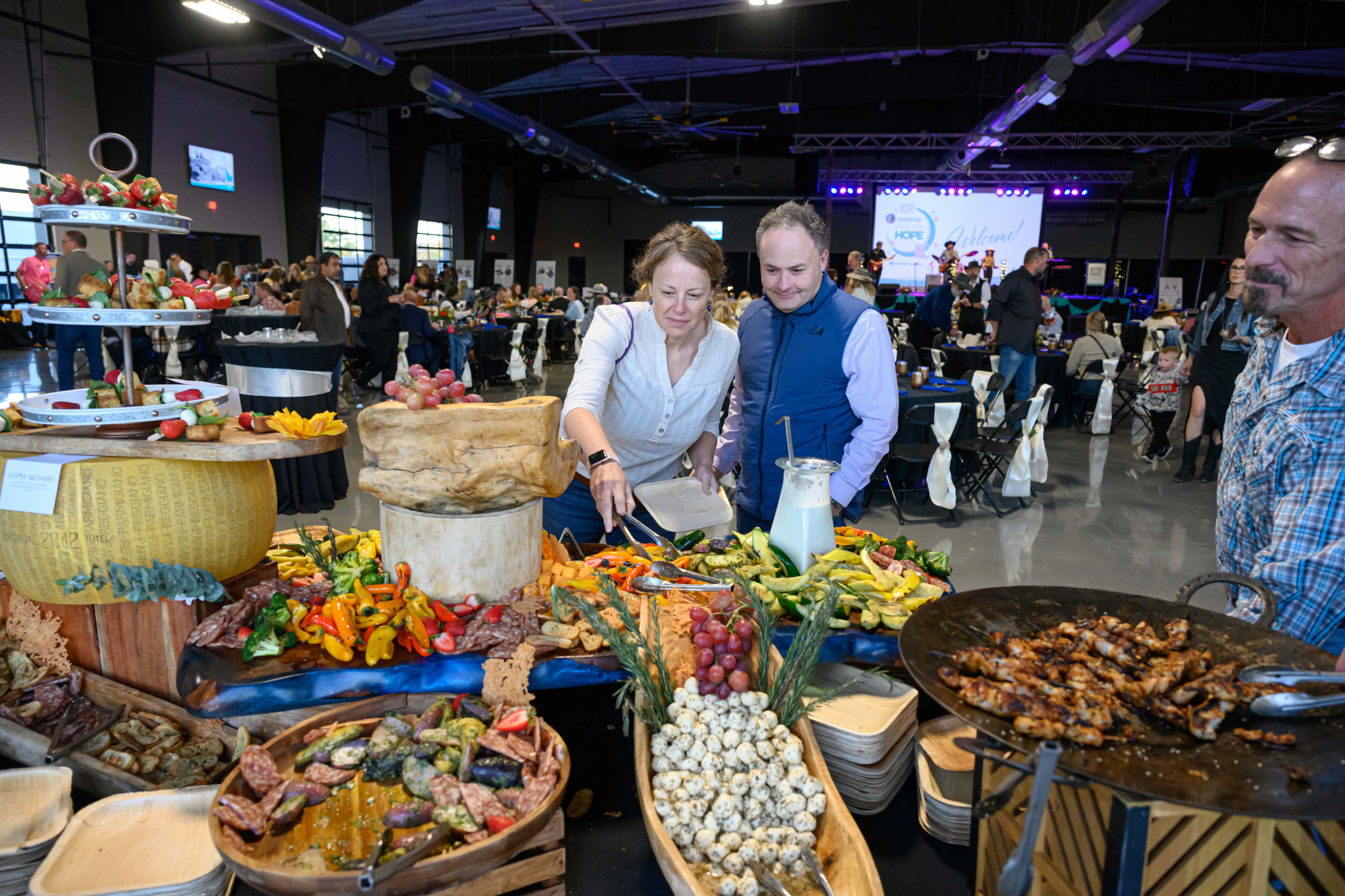 People serving themselves food from a buffet with vegetables, cheese, and grilled meat at an indoor event.