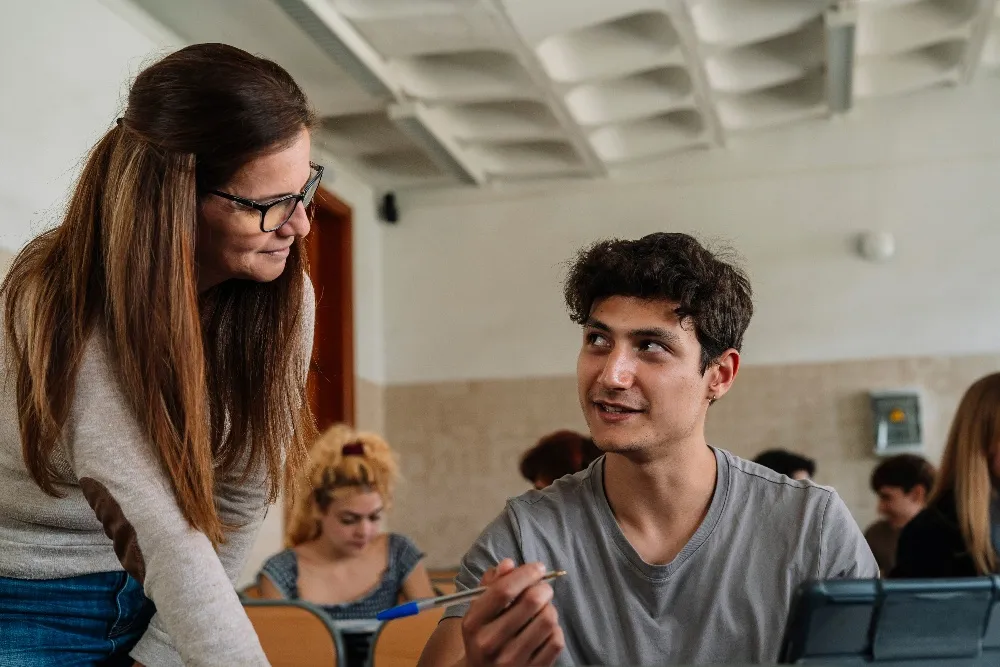 Teacher with glasses leaning over to help a male student holding a pen in a classroom.