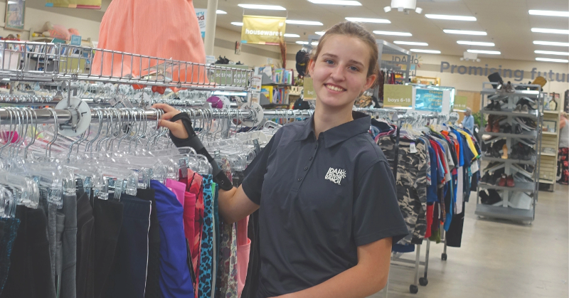 Smiling youth wearing an Idaho Youth Ranch shirt standing by clothing racks in a store.
