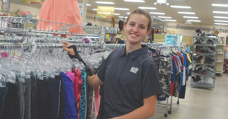 Smiling youth wearing an Idaho Youth Ranch shirt standing by clothing racks in a store.