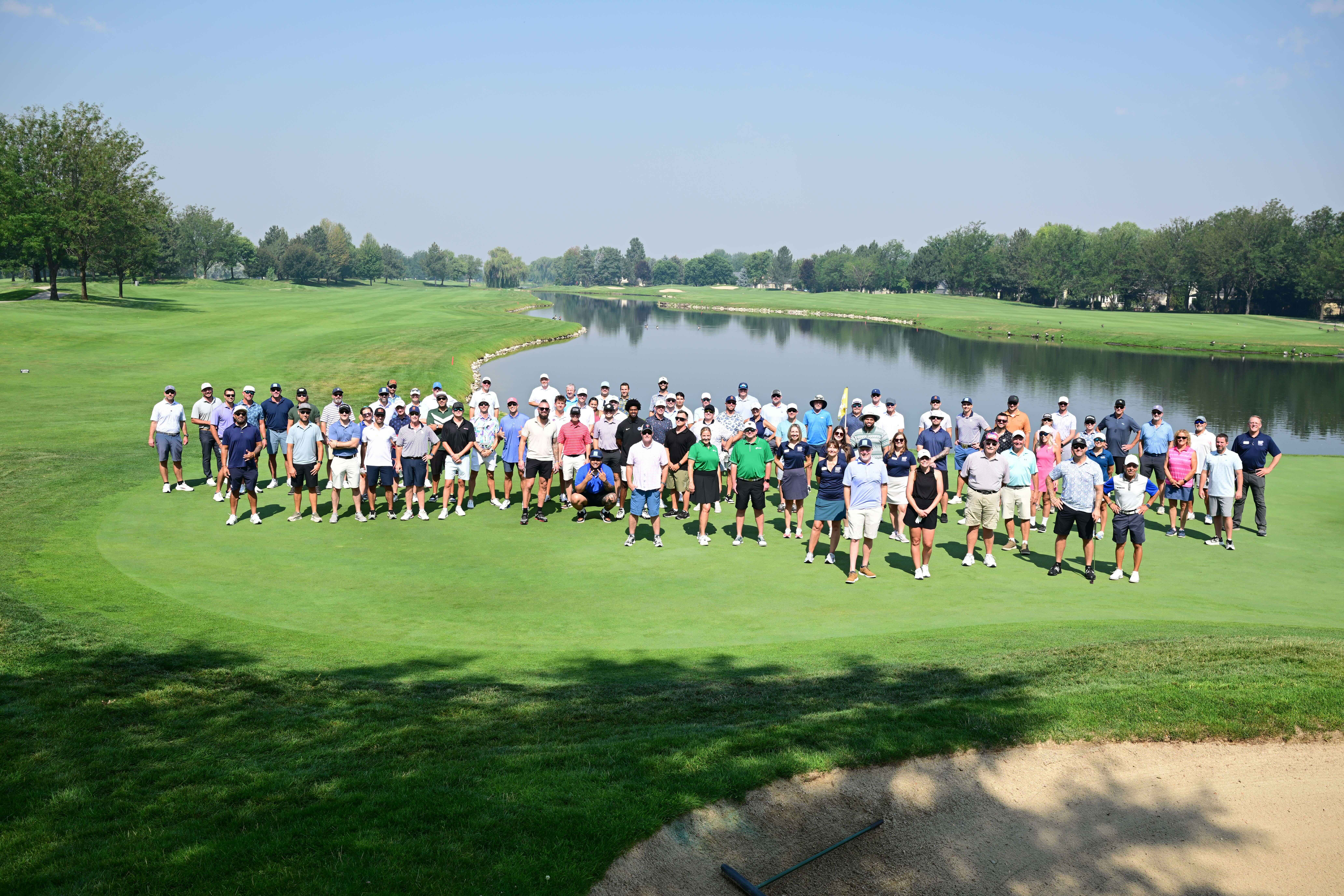 Group of people standing on a golf green by a lake under a clear blue sky.