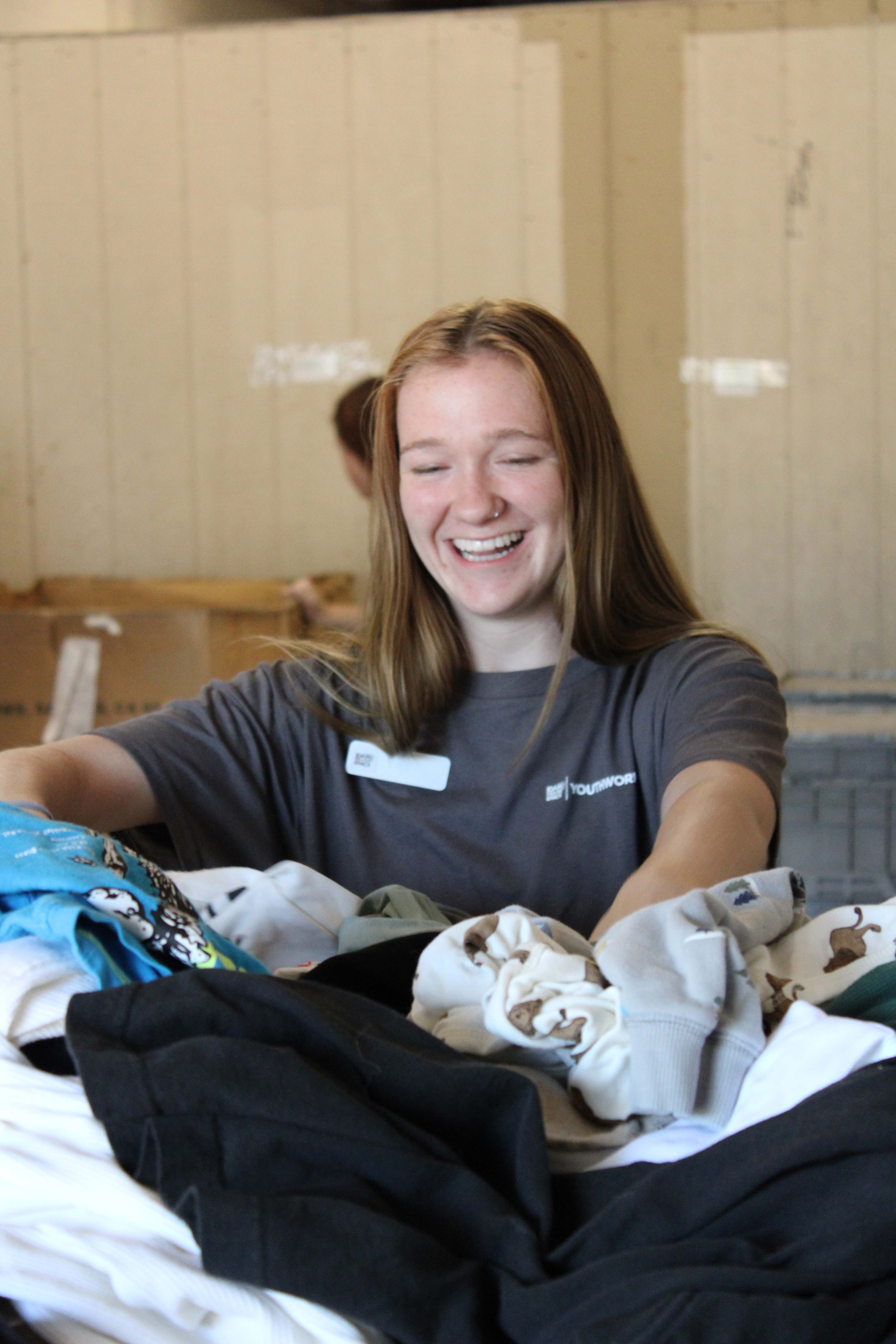 Young woman in gray shirt smiling while sorting a pile of clothes indoors.