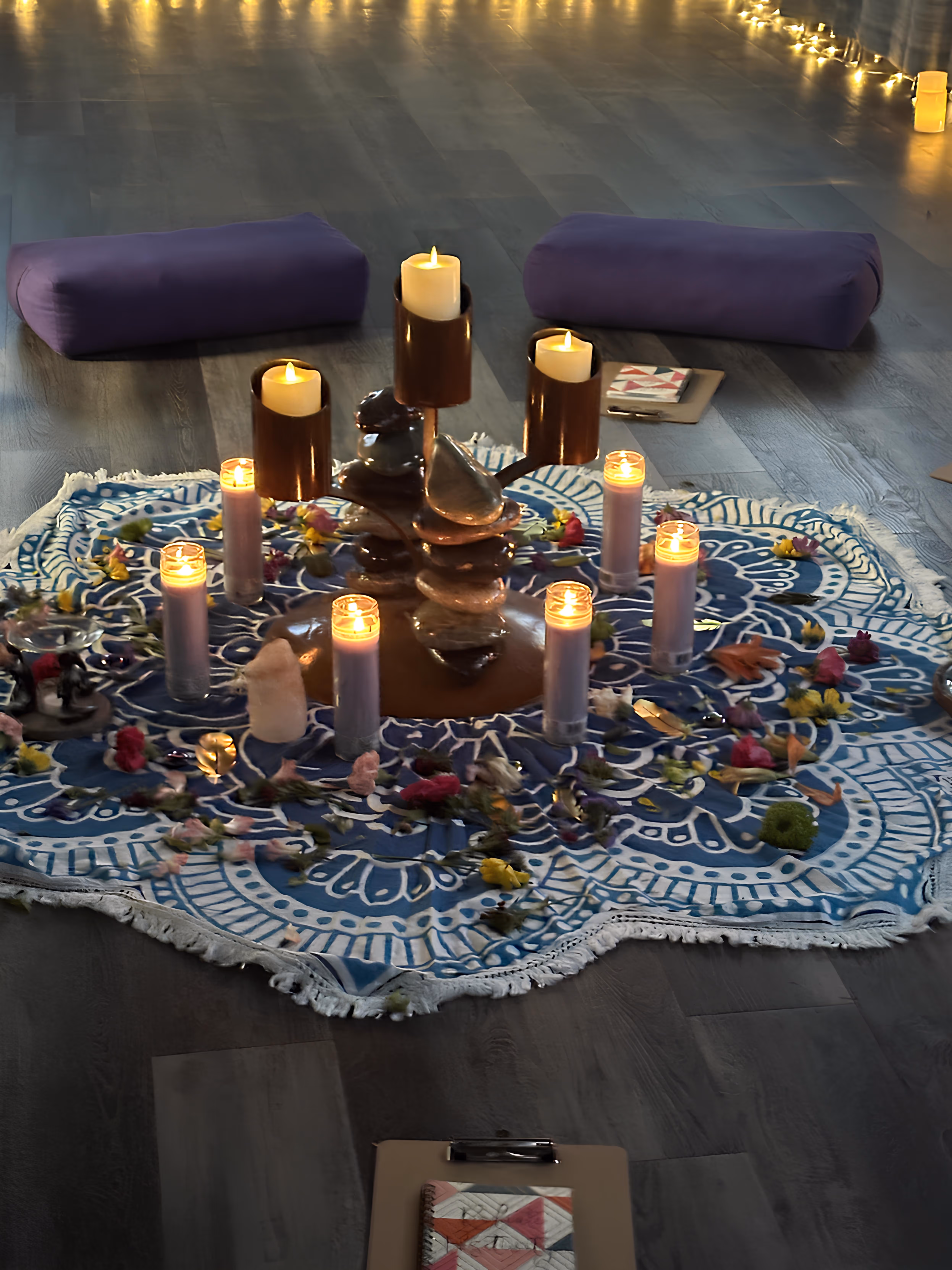 Meditation setup with lit candles, flower petals, stones on a blue patterned cloth, and two purple cushions on a wooden floor.