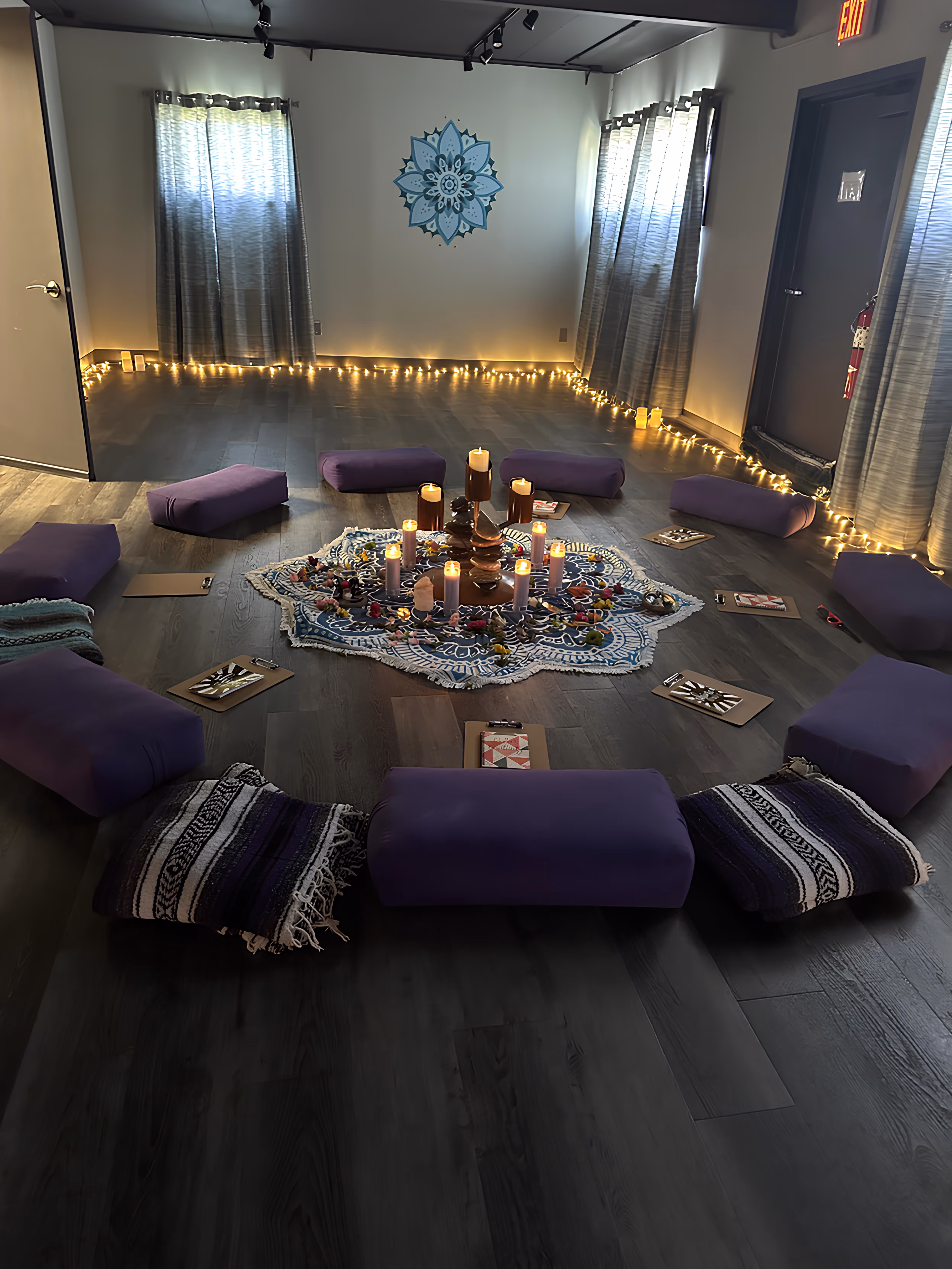 A meditation room setup with purple cushions and blankets arranged in a circle around a decorative centerpiece with lit candles on a patterned cloth.