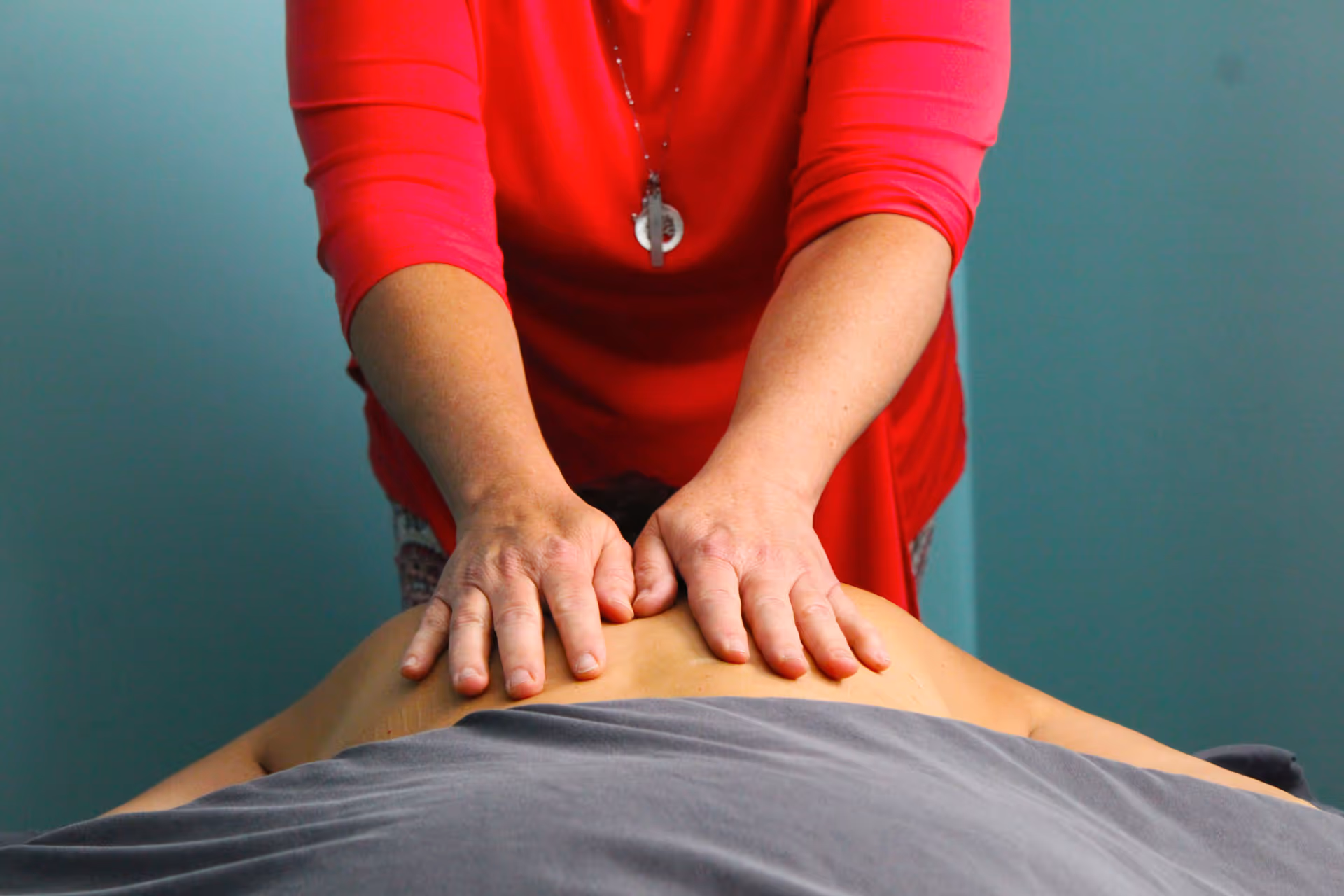Person in red shirt giving a back massage to another person lying face down covered with a gray sheet.