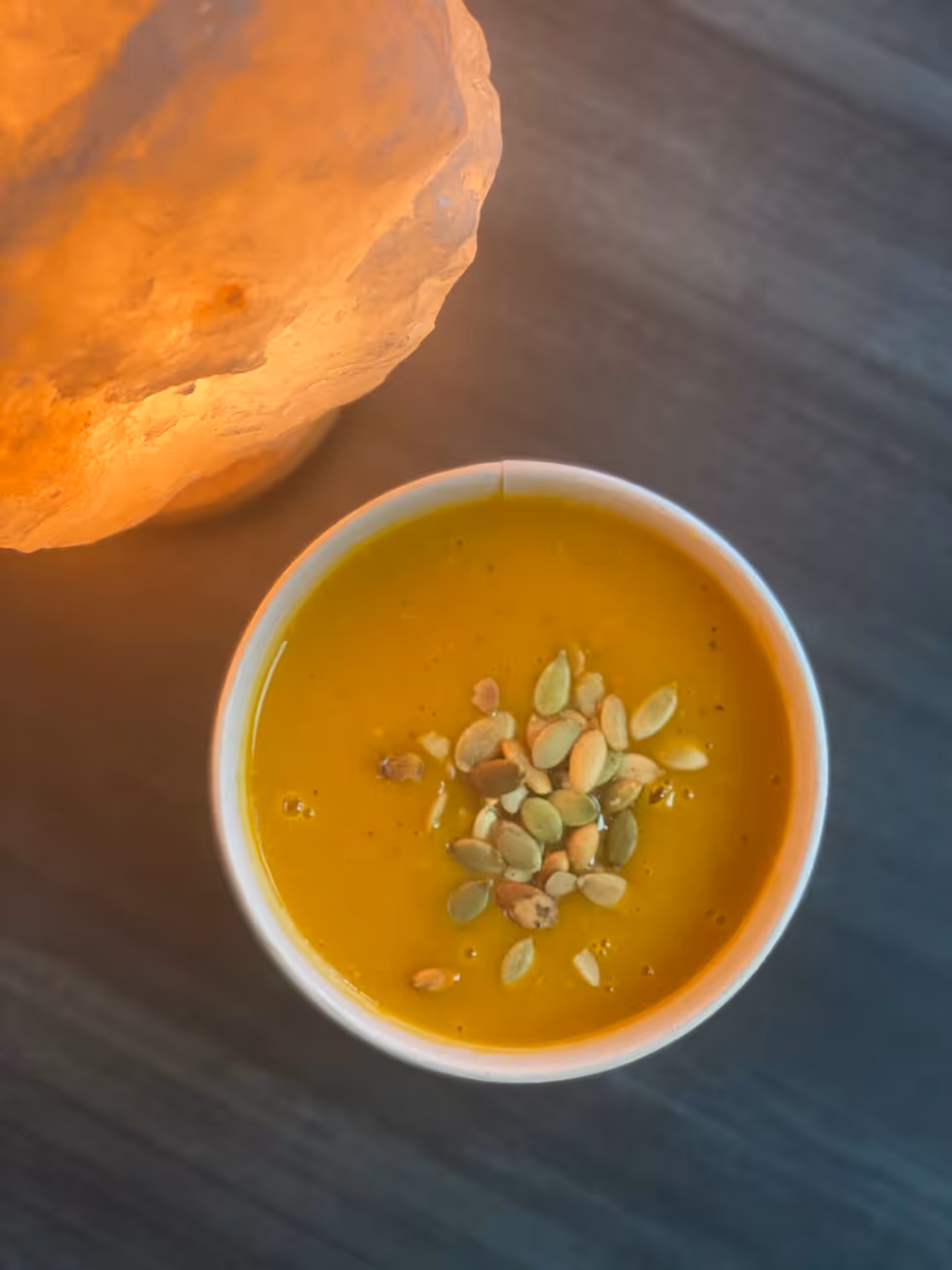 Bowl of creamy orange soup garnished with pumpkin seeds on a dark surface next to a glowing salt lamp.