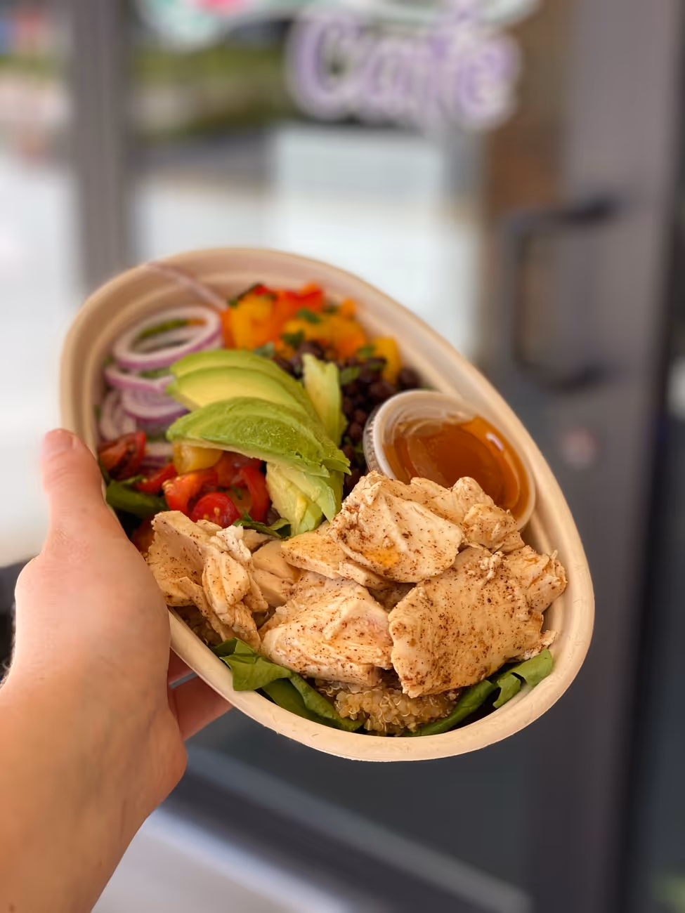 Hand holding a bowl with grilled chicken, avocado slices, red onion, tomatoes, black beans, spinach, quinoa, and a small container of dressing.