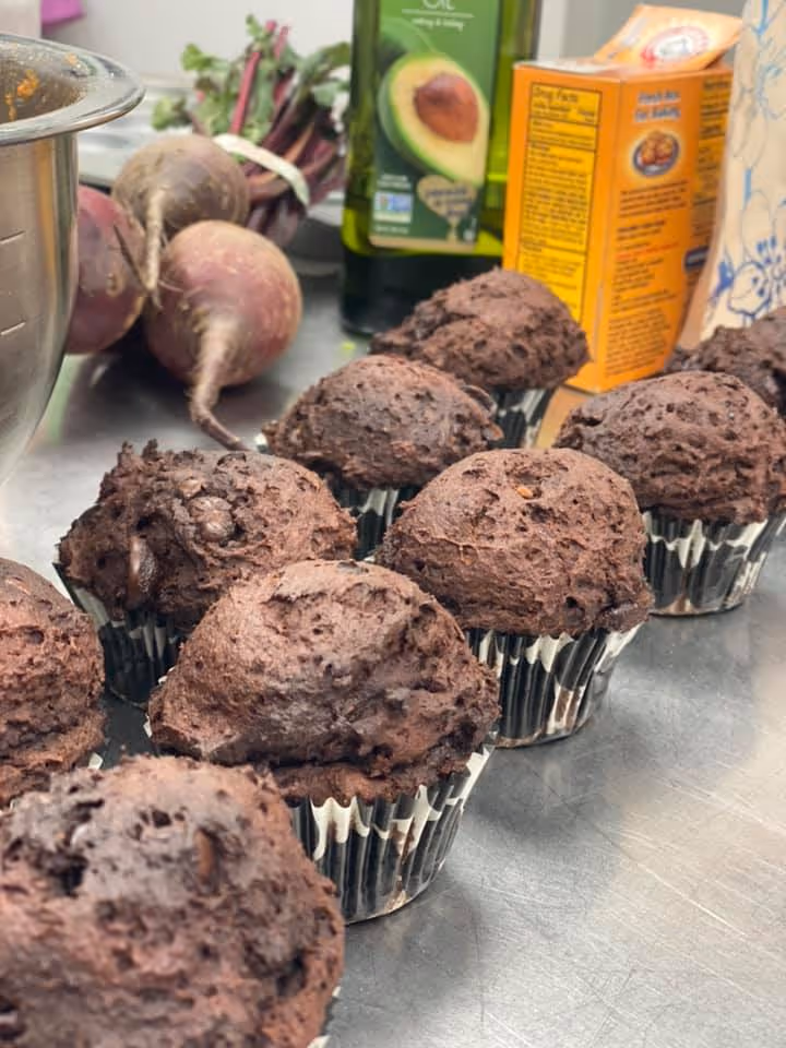 Row of chocolate muffins on a kitchen counter with fresh beets, an olive oil bottle, and a baking mix box in the background.
