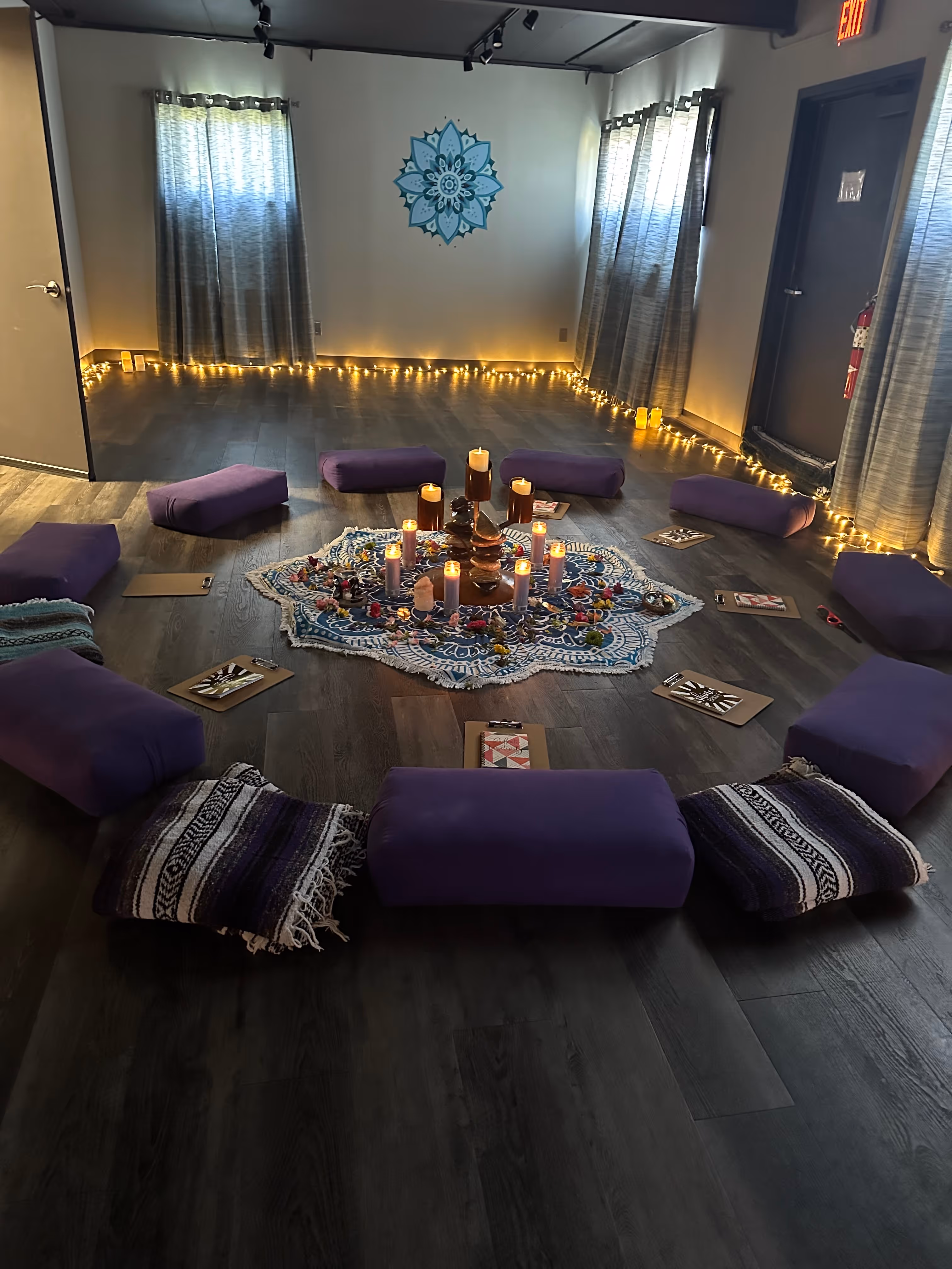 Meditation room with purple cushions and striped blankets arranged in a circle around a central area decorated with lit candles and flowers on a patterned cloth.