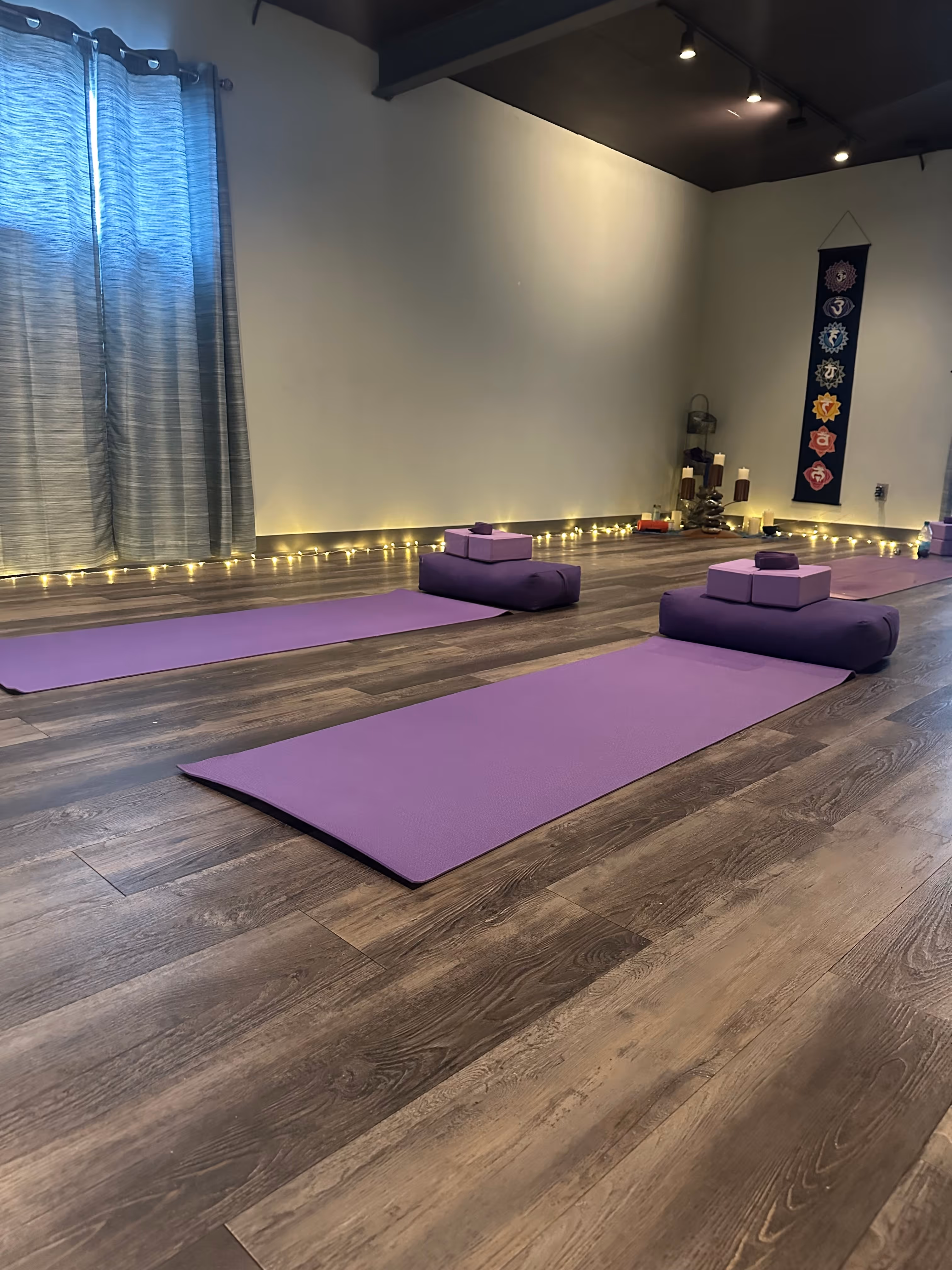 Yoga studio with two purple mats each topped with a purple bolster and yoga blocks, soft lighting, and a chakra wall hanging.