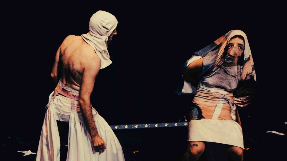 Two performers, one hooded, mirroring each other on a dark stage