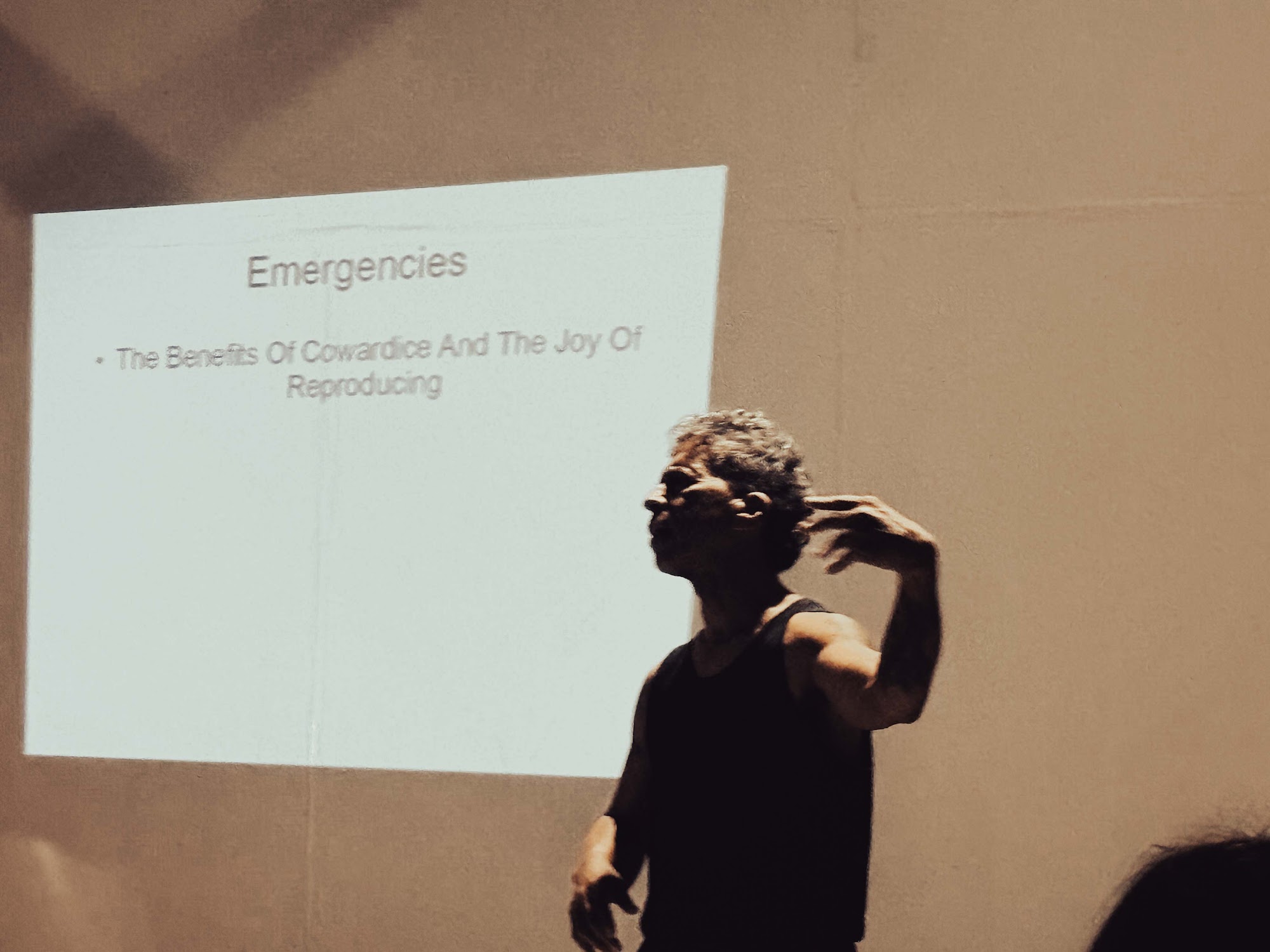 Michael Gougis uses expressive hand gestures while discussing a slide titled 'Emergencies' in a performance space.