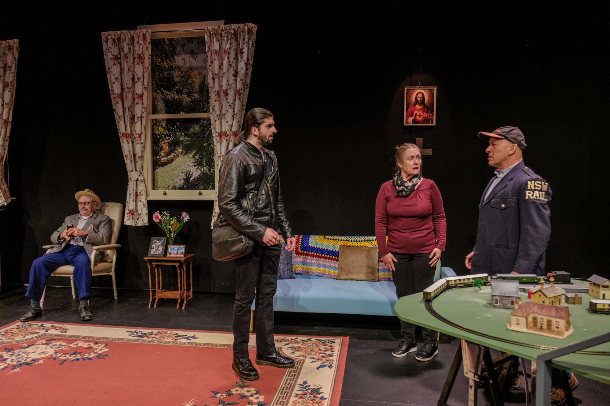 A mother and son stand facing each other in an indoor living room while a grandfather sits in the background.