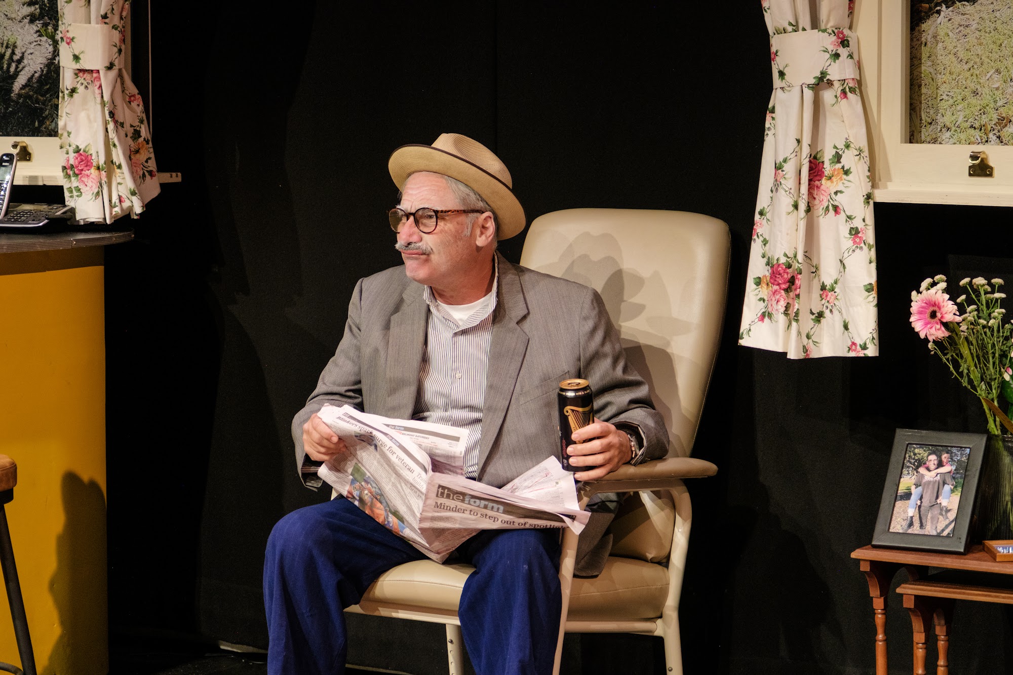 A man sits in a chair reading a newspaper, holding a drink, while appearing relaxed.