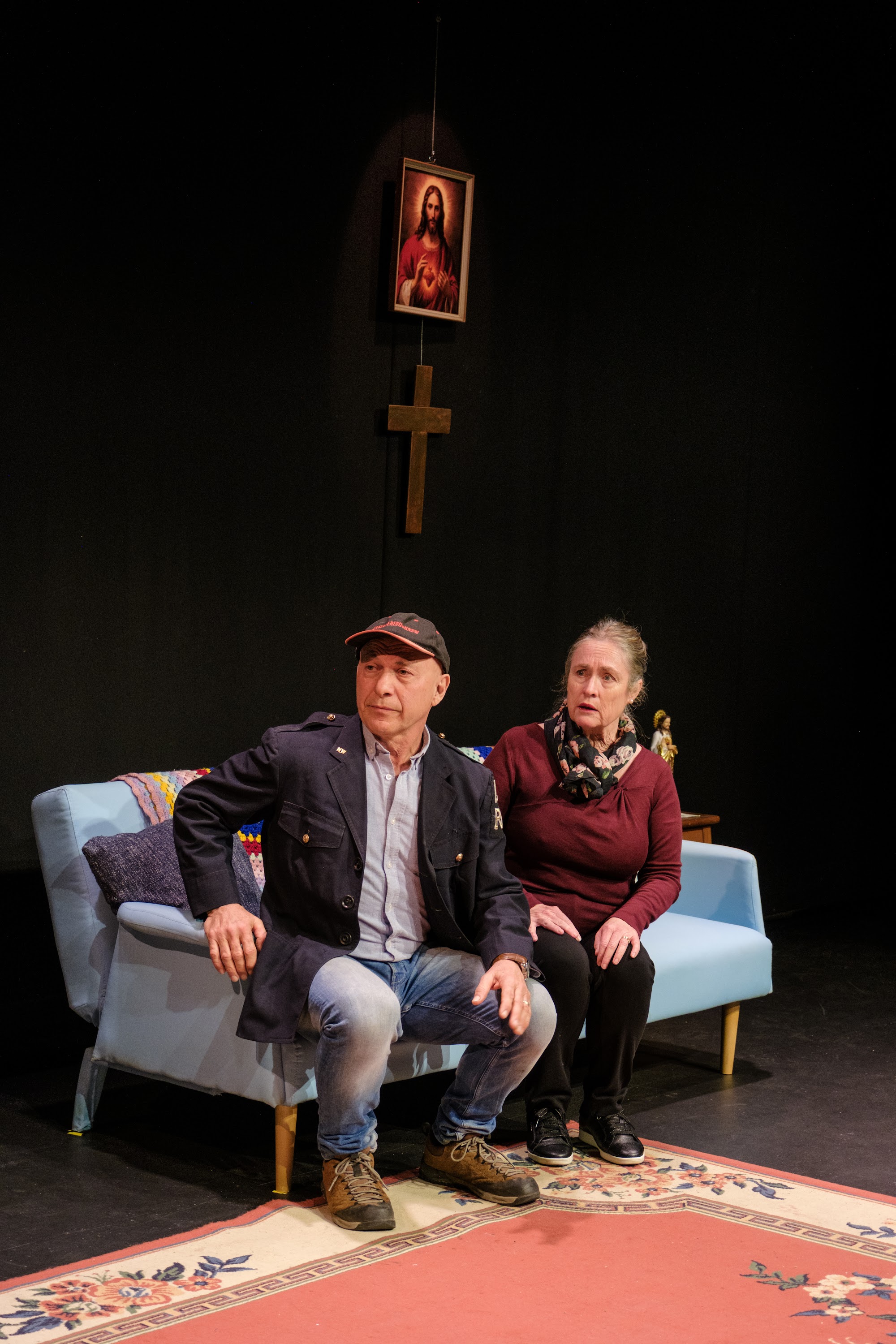 A mother and grandfather sit on a couch, both looking concerned while a cross and Jesus painting hang above them.