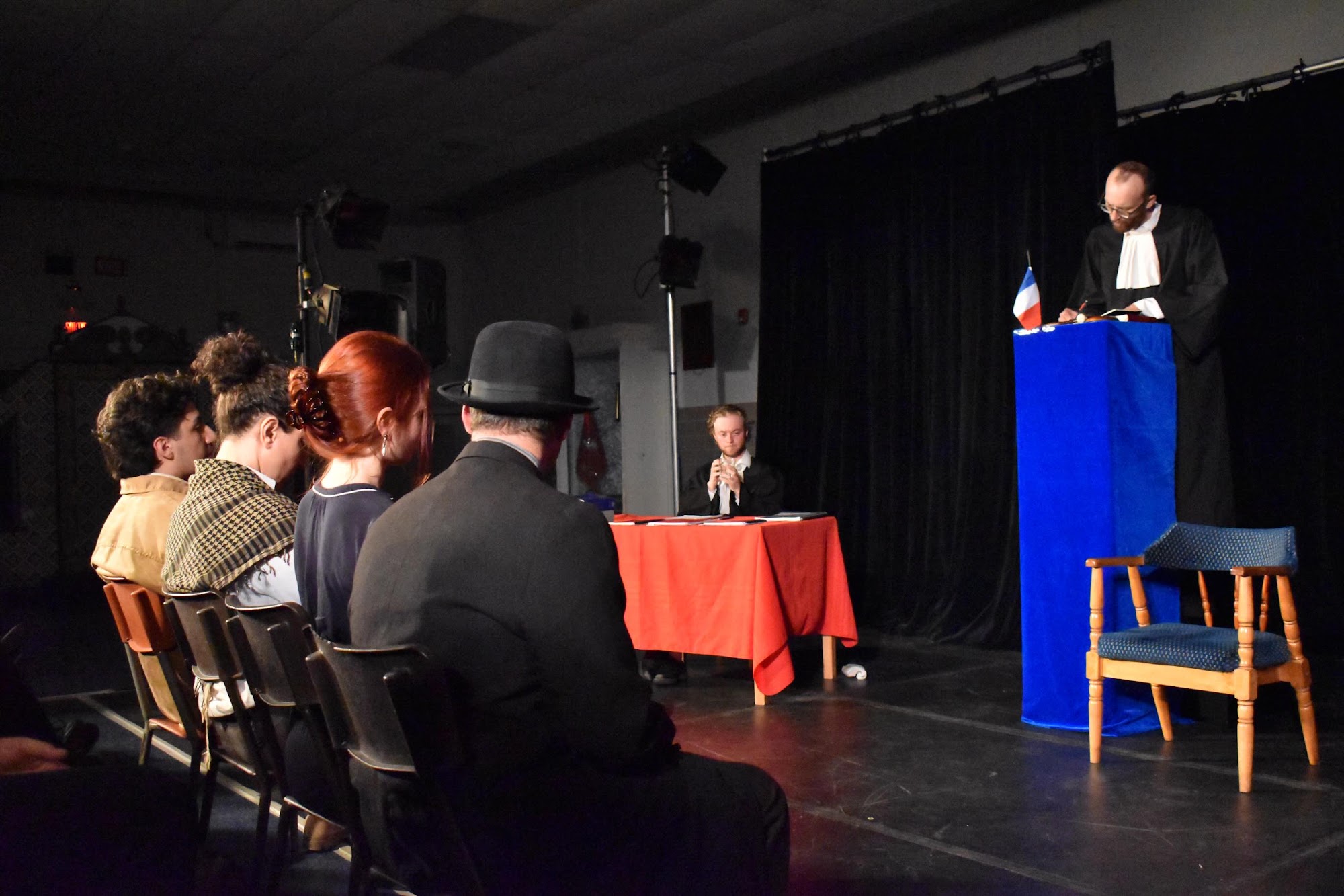 A wide view from behind shows rows of seated spectators facing a lawyer at a podium on a dimly lit stage, with an empty chair beside him.