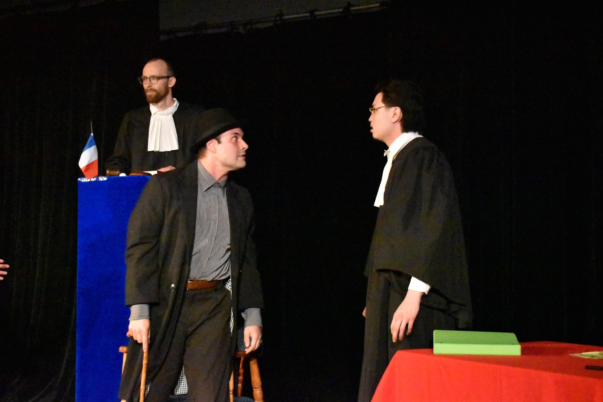 A lawyer in black robes stands in front of a man in a bowler hat and long coat, engaged in tense conversation on a dimly lit stage.