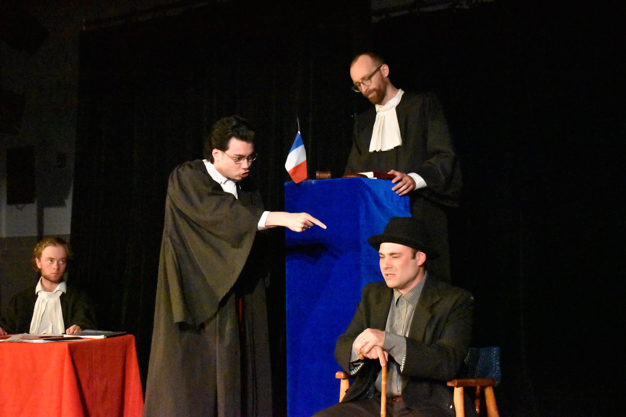 A lawyer in black robes points accusingly toward a podium while another man in a bowler hat sits nearby looking down; a second lawyer and other seated figure are visible in the background.
