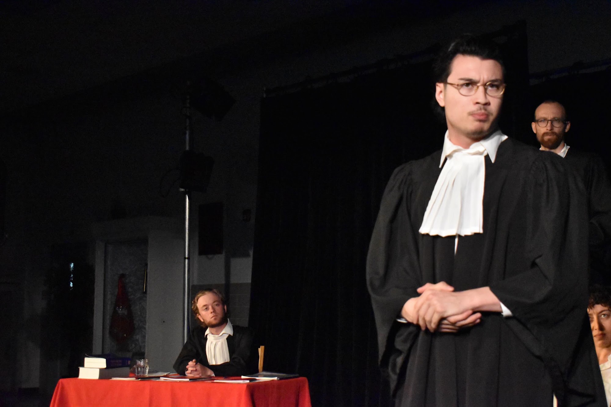A close-up of a lawyer in glasses and a white cravat with a stern expression, another lawyer visible blurred in the background.