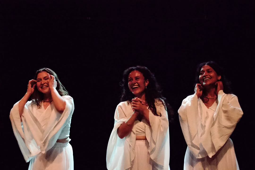 Three dancers in white robes stand side by side on a dark stage, laughing together after a performance.