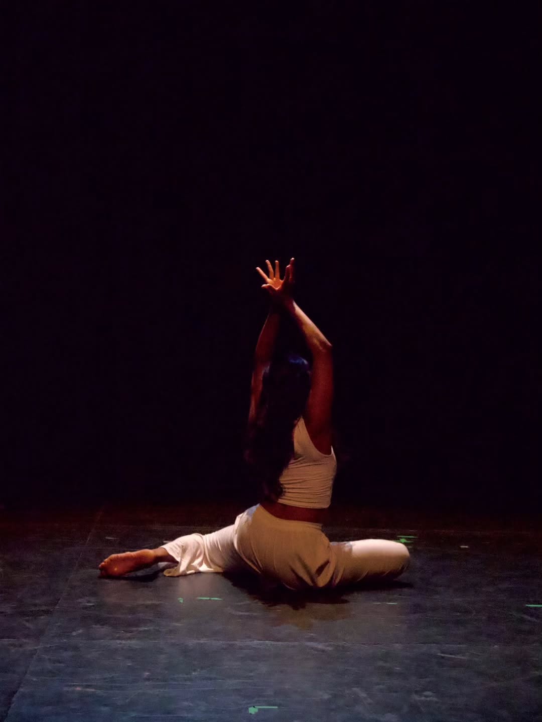 A dancer in cream-toned clothing kneels on stage with her back to the audience, arms raised overhead in a moment of stillness.