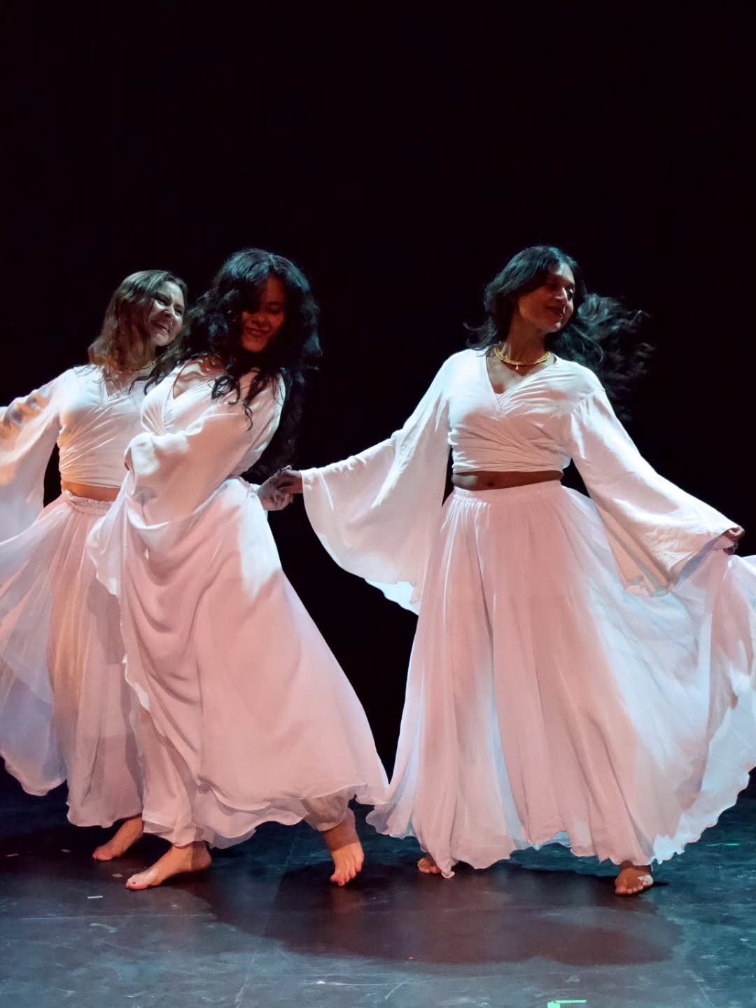 Three dancers in flowing white skirts move together across a dark stage, hands linked, fabric swirling.