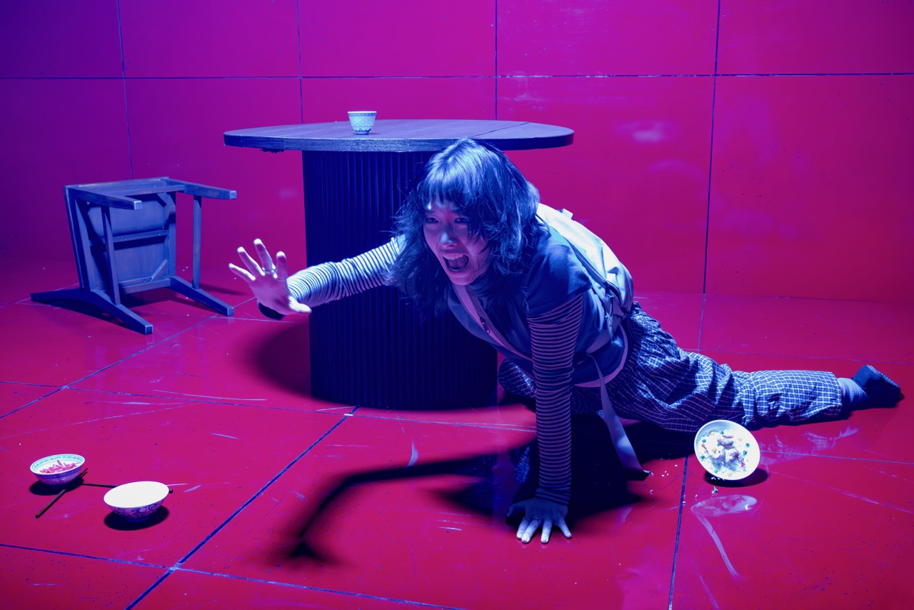 Claudia crawls on the floor, reaching out in desperation, with a knocked-over chair and spilled food nearby.
