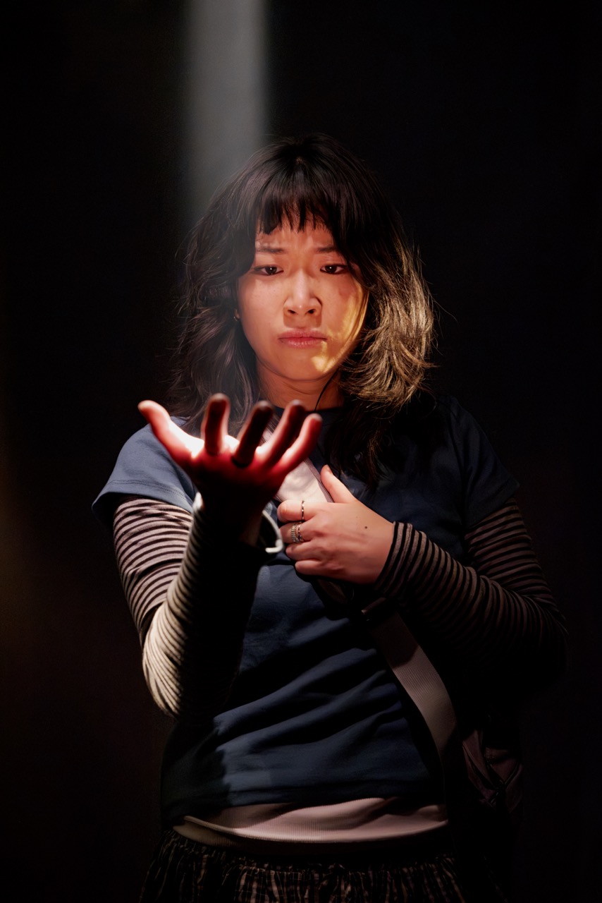 Claudia stands in a focused pose with one hand extended, illuminated by a spotlight.
