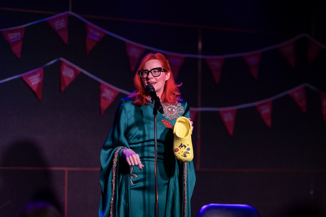 Glynis Traill-Nash on stage in a green gown, holding a decorated yellow shoe.