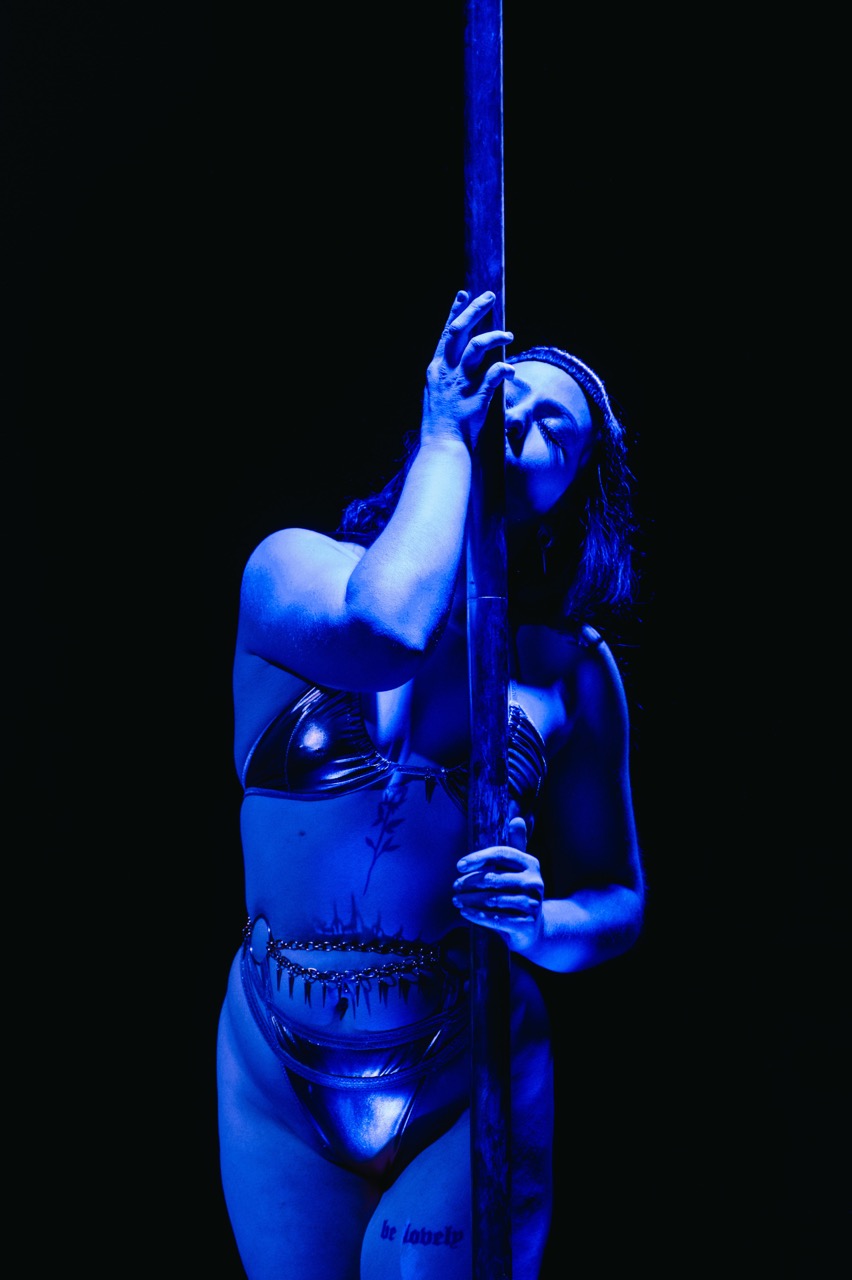 Cleo Rapture in a metallic bikini leans against a pole under blue stage lights.