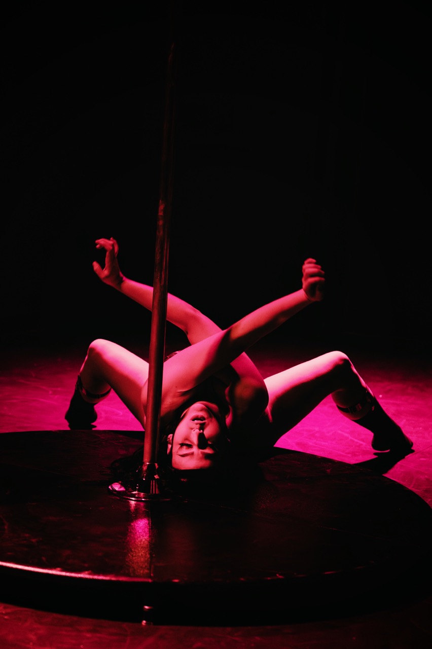 Cleo Rapture lies inverted on the stage floor, gripping the pole with both hands, legs stretched wide.