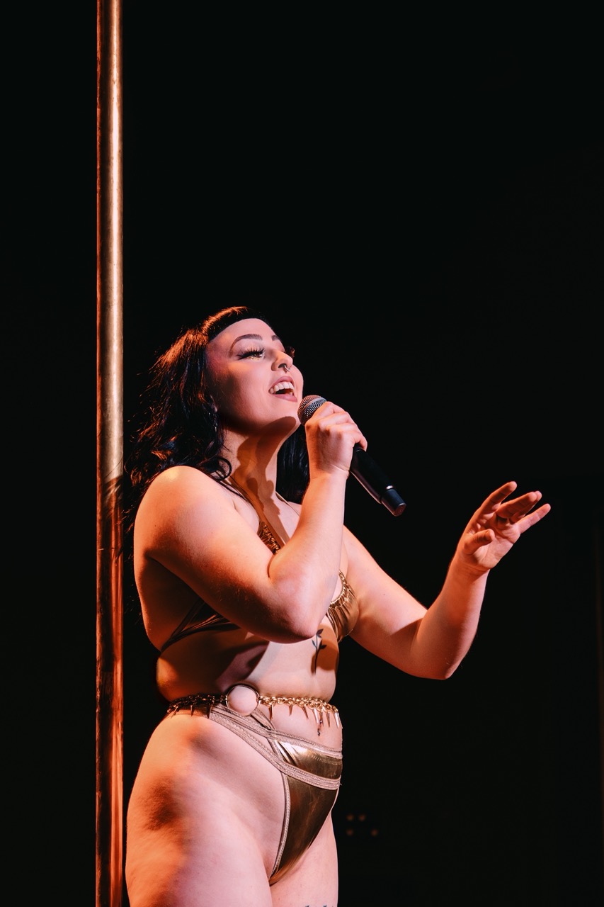 Cleo Rapture holds a microphone and gestures animatedly to the audience.