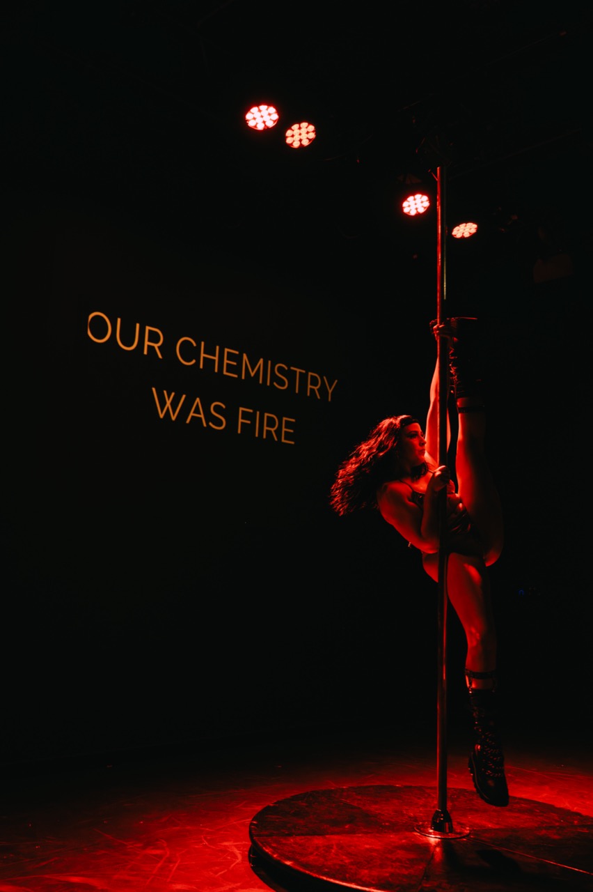 Cleo Rapture poses on the pole with one leg extended, a projected caption reading 'Our chemistry was fire' behind her.