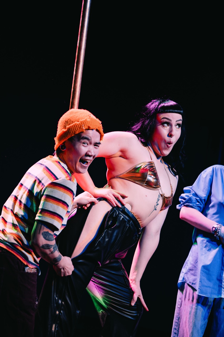 Cleo Rapture reacts with hands on hips while a person in an orange beanie playfully leans in beside her.