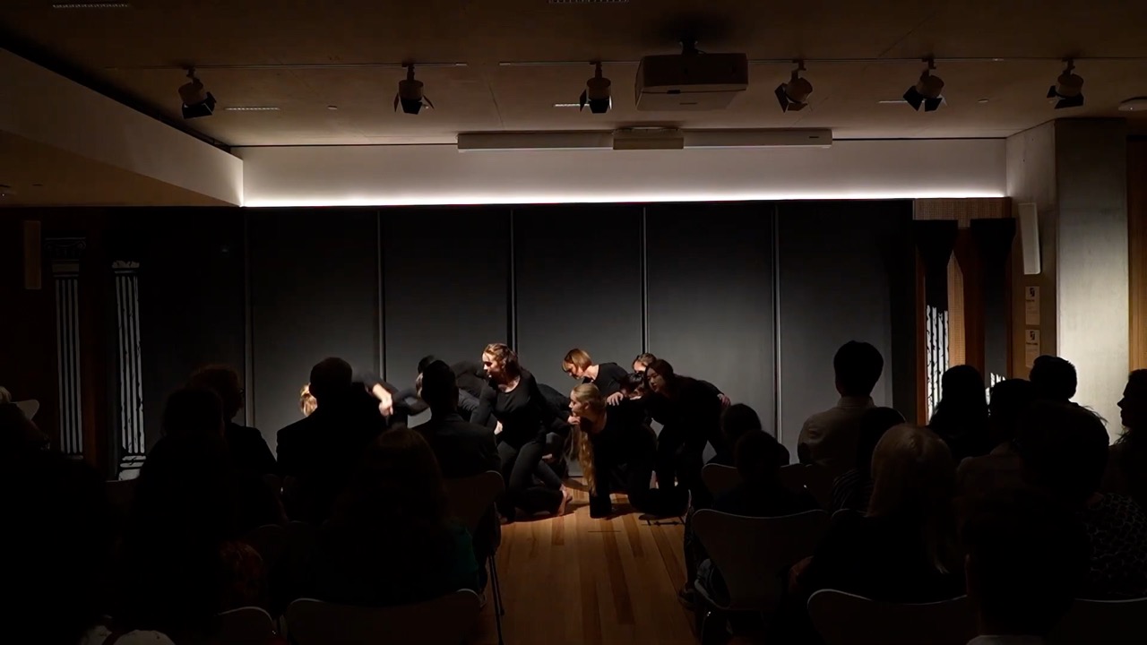 A group of students and academics gathered around a large wooden table. Photographs and materials are spread across the surface. Bookshelves line the wall behind them; one person leans in to explain something to the group.