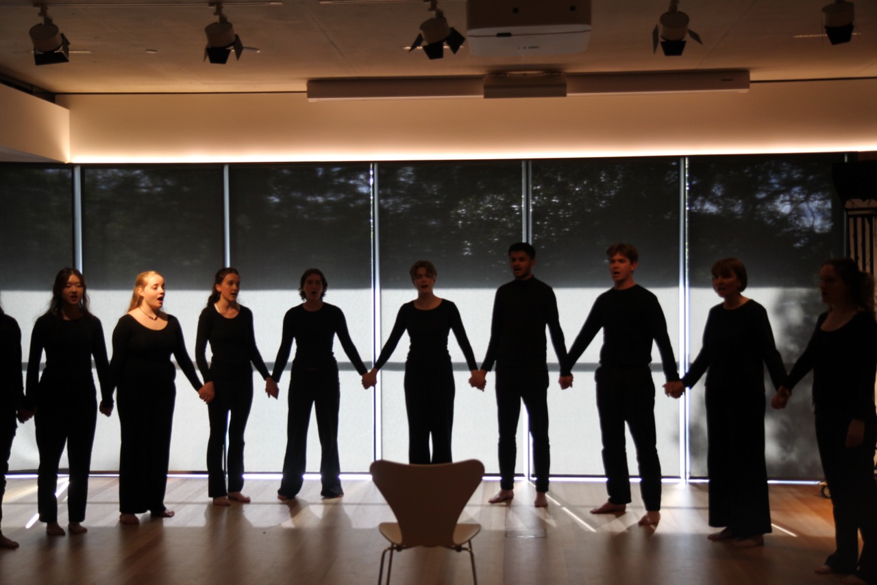 Ten chorus members dressed in black stand in a straight line across the stage, holding hands. An empty modernist chair sits at centre front. Translucent window blinds form the backdrop, with faint daylight showing through.