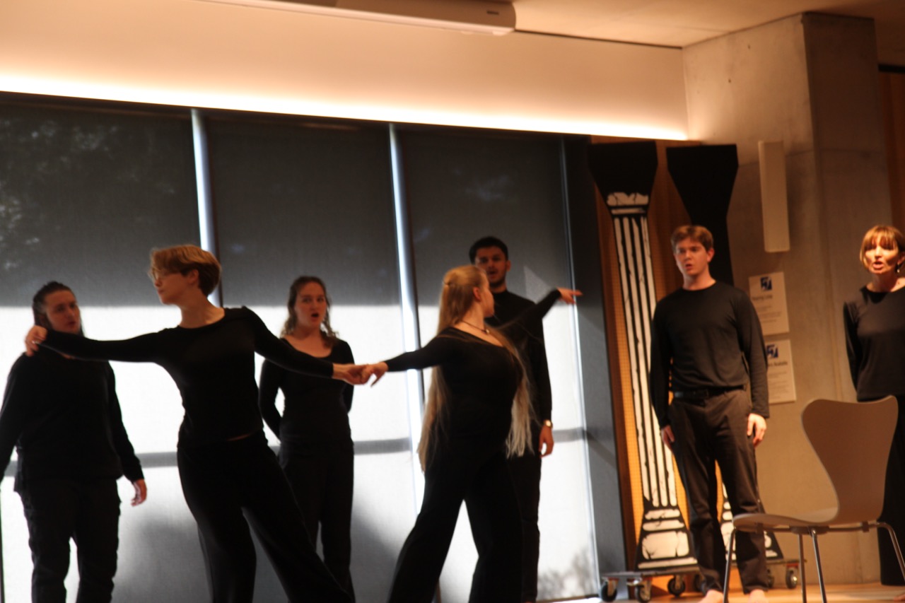 A darkened performance space. The chorus is clustered together at centre in low light, seen over the heads of audience members silhouetted in the foreground.