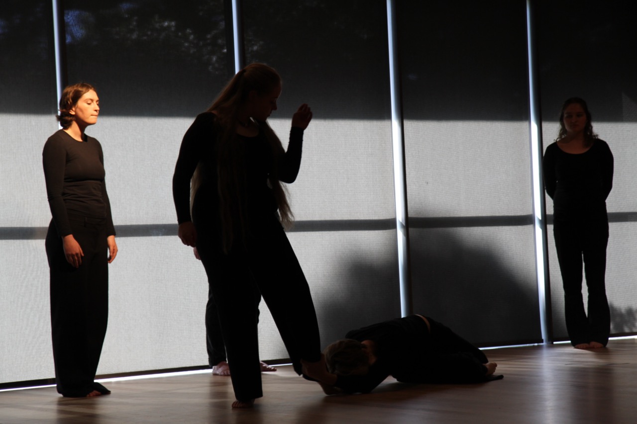 Chorus members in black dance with arms extended, one figure caught mid-turn with hair swinging. A classical column set piece stands at right, beside a still figure watching.