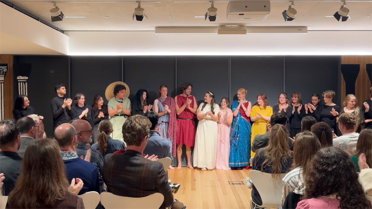 Five actors pose casually in Greek costume. A woman in cream stands at left, then a woman in pink; a man in a blue chiton and red drape stands at centre wearing a headband; another man in blue with a striped drape stands beside him; a woman in a red-and-blue robe stands at right. A dark-clothed figure passes in the background.