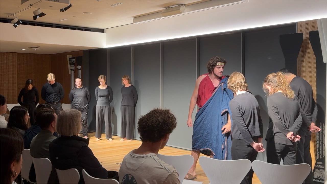 Four figures on a starkly lit stage. One woman stands at left with her eyes closed; another figure gestures with a raised arm at centre; a third has collapsed onto the floor; a fourth stands watching at right.