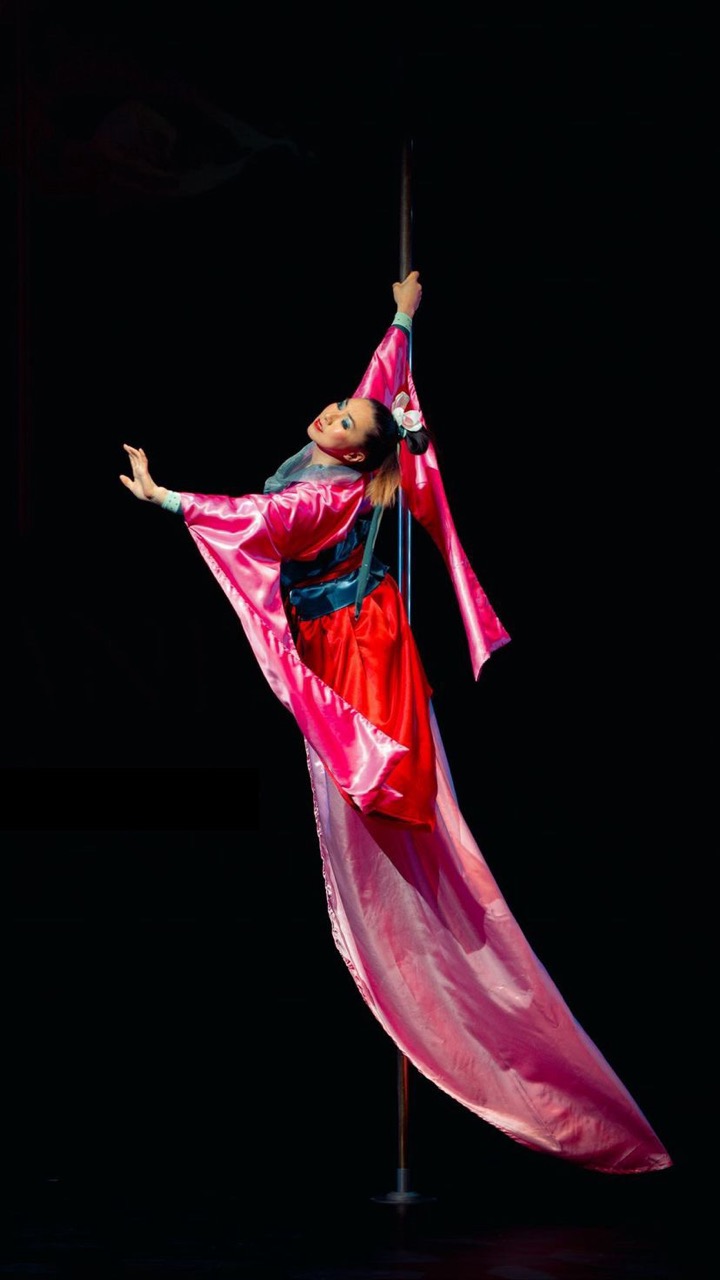 Andrea James Lui balanced on a pole in a flowing pink and red costume, arms outstretched, gazing toward the audience.