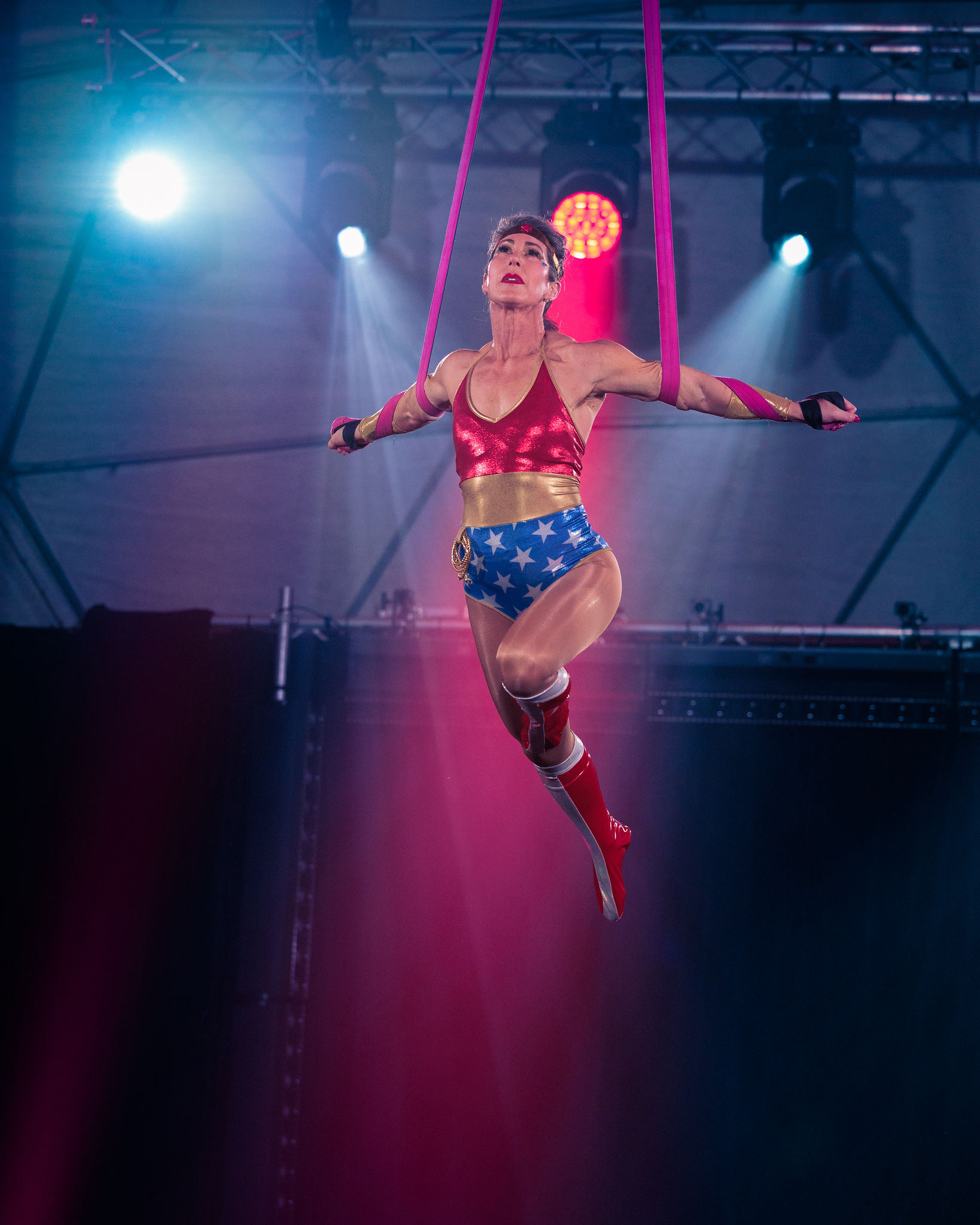 Hazel Palmer in a superhero costume swings from pink aerial silks, one arm extended toward the audience.