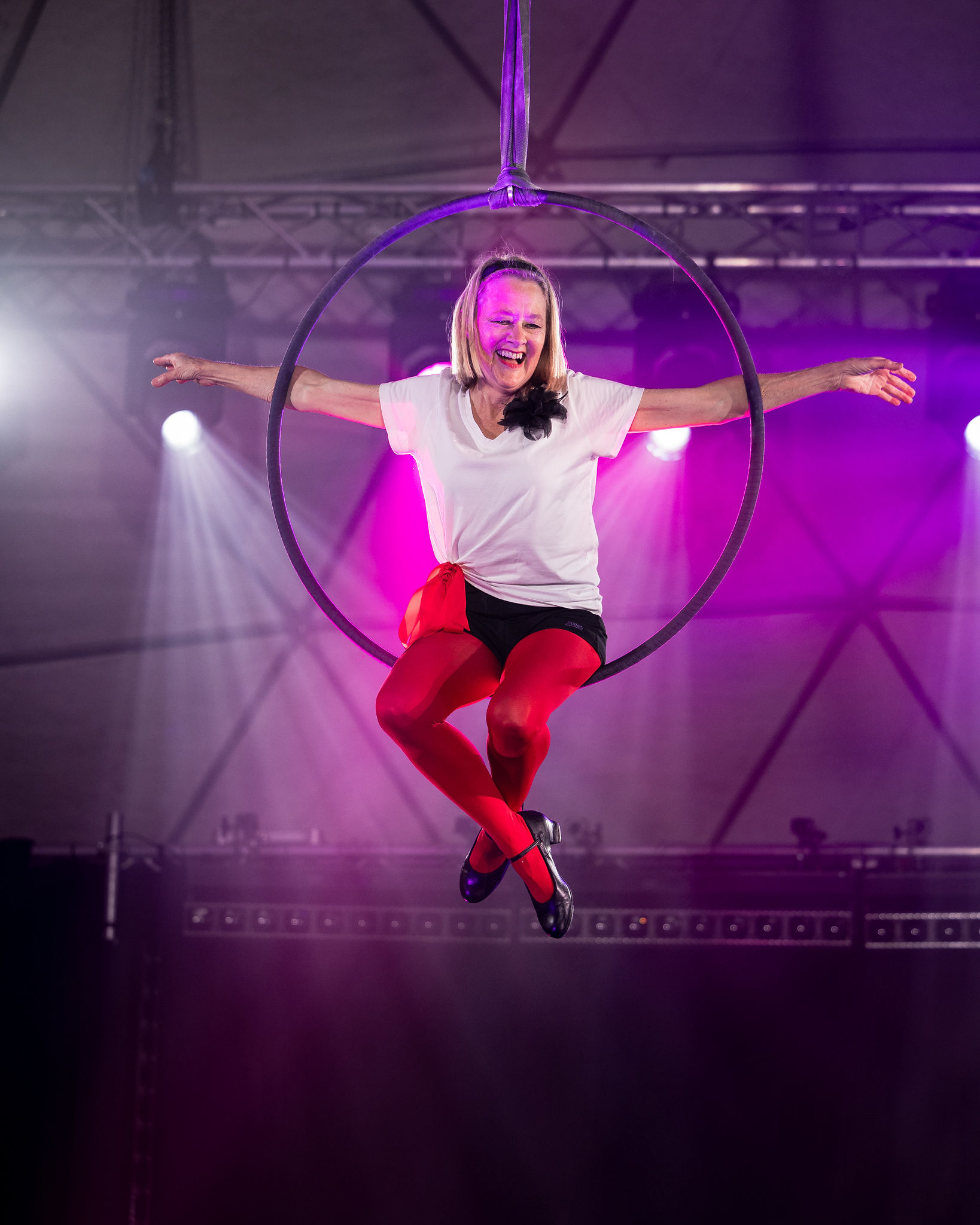 A woman in a white shirt and red tights smiles widely as she sits in an aerial hoop above the stage.