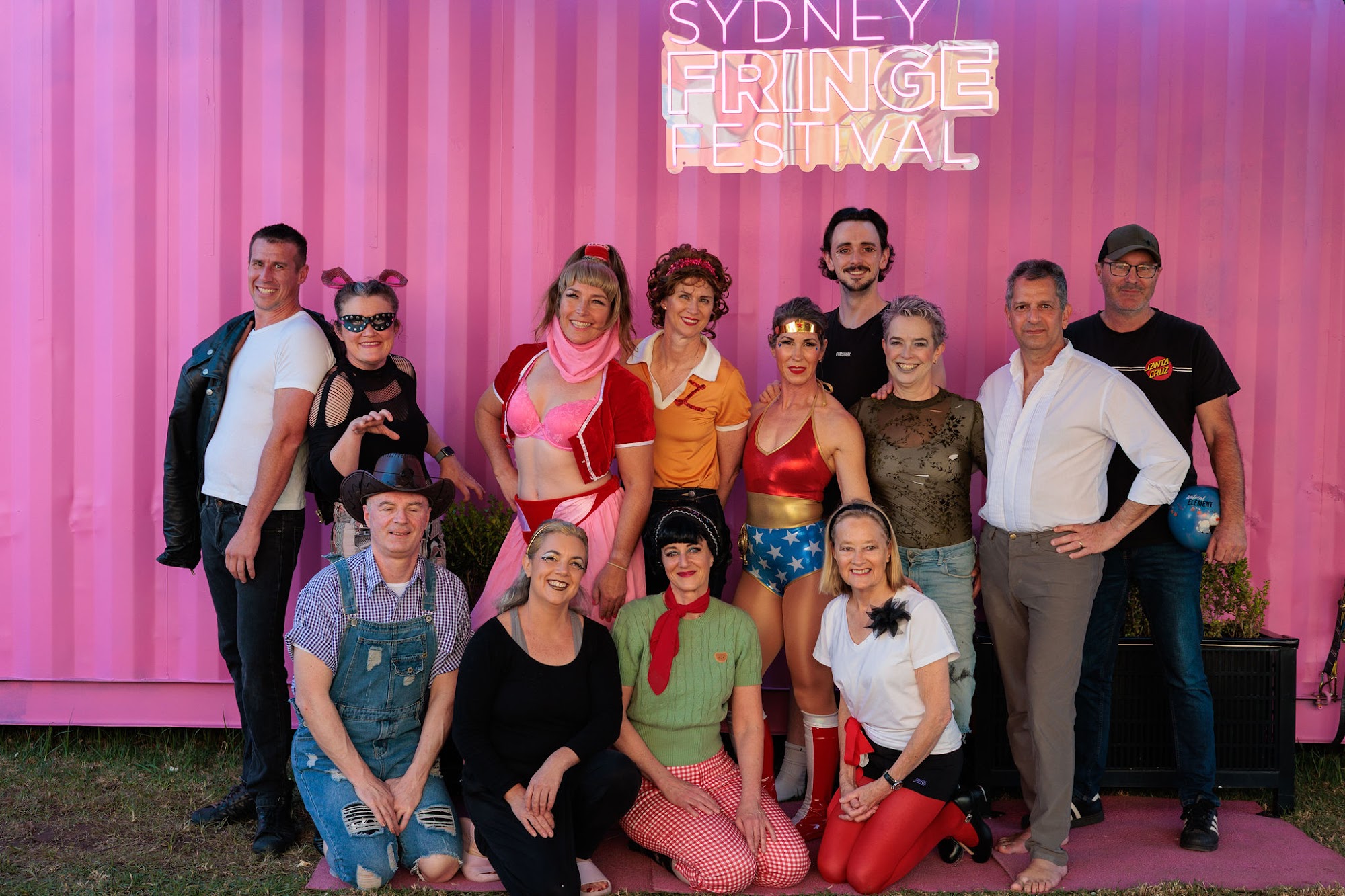 A diverse group of 50-Up performers pose in colourful retro costumes in front of a pink Sydney Fringe Festival backdrop.