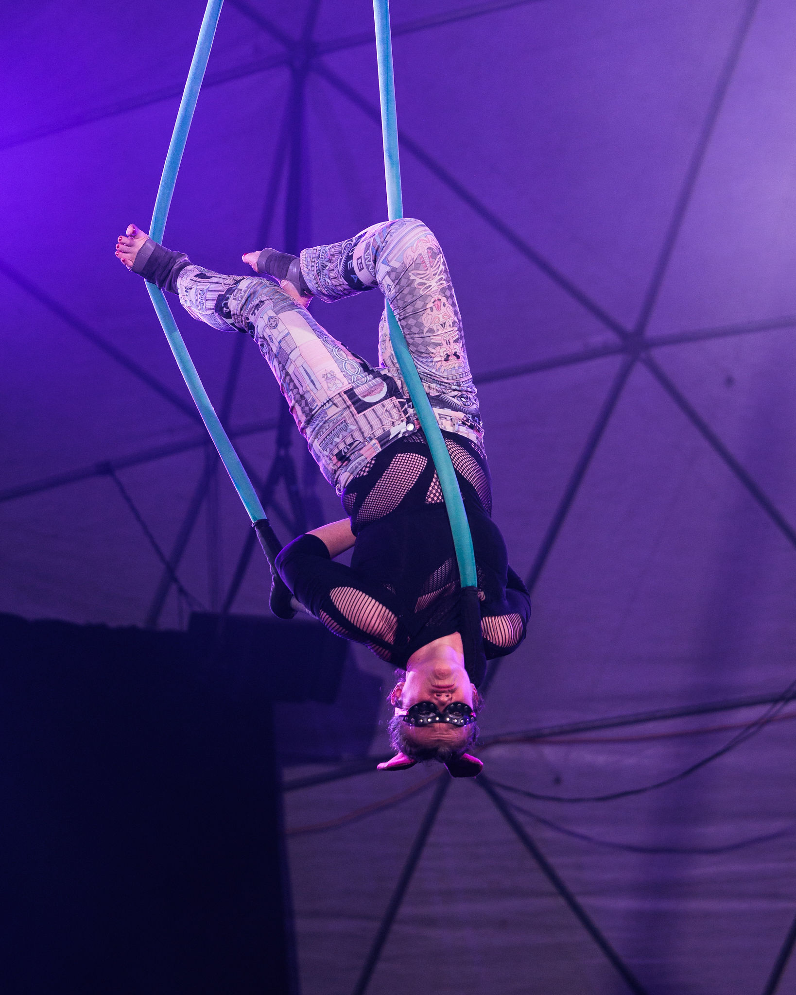 An aerialist hangs upside down from blue silks with legs crossed, gripping the fabric tightly.
