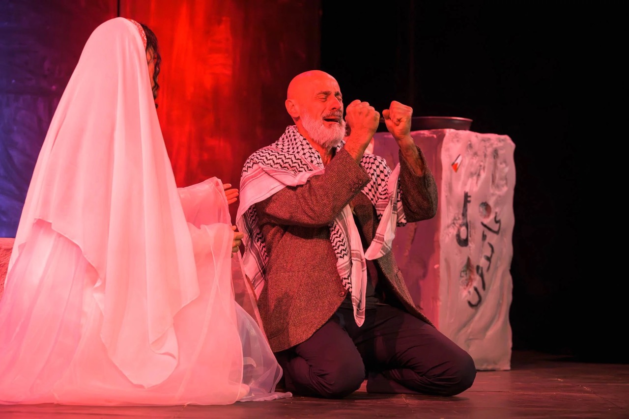 A man kneels on the stage with clenched fists, while a woman stands beside him.