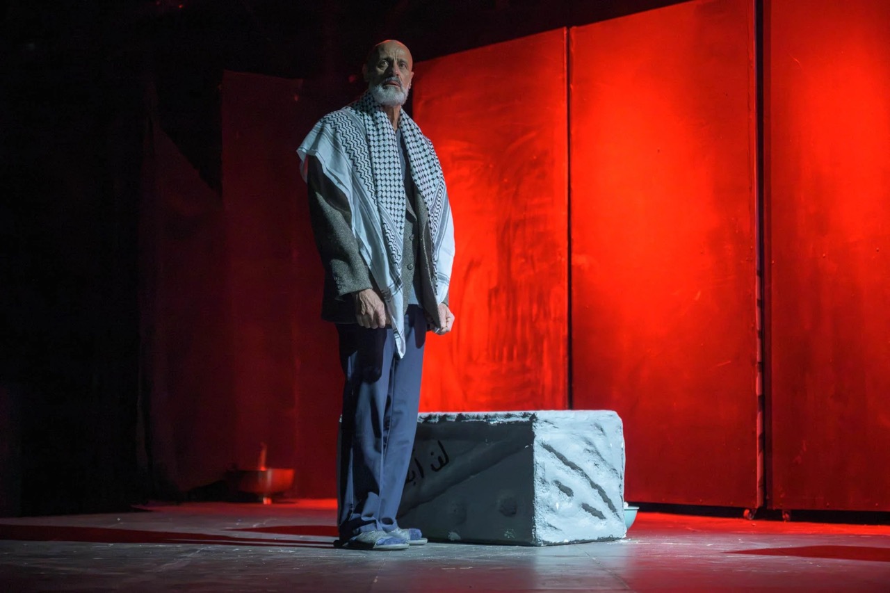 A man stands alone on stage beside a small stone platform, looking out into the audience.