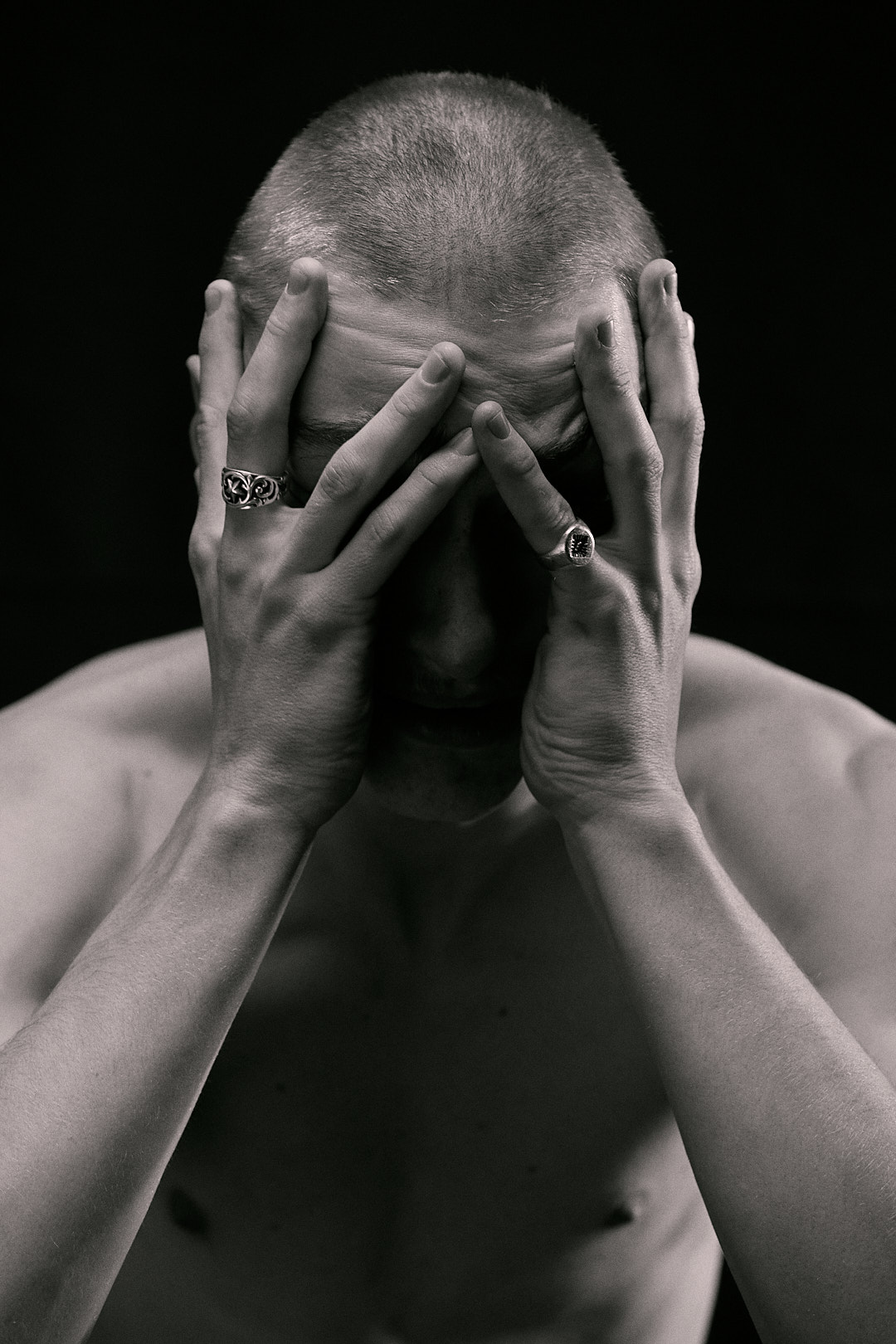 A black and white image of Jake Davies shirtless, with his head buried in his hands. He wears a ring on his left pinkie and right ring finger.